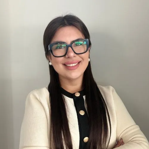 Smiling portrait of a woman with long dark hair and black-framed glasses, wearing a cream cardigan over a black top with gold buttons.