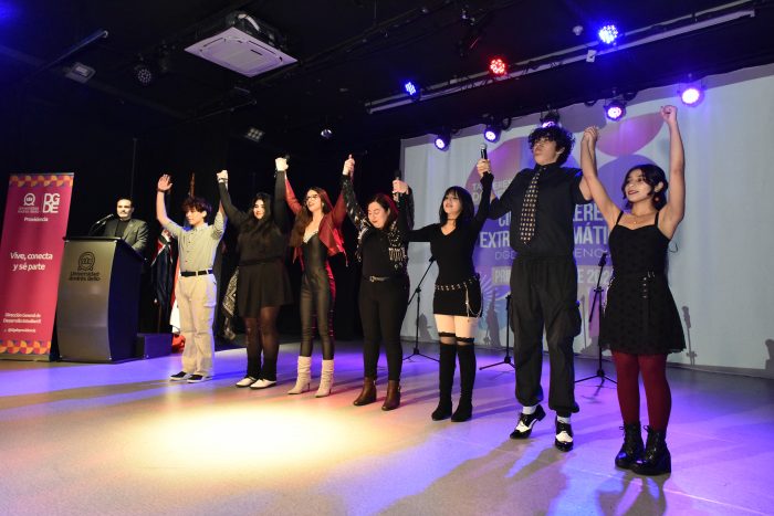 Group of performers on stage, holding hands up in triumph, facing audience under stage lights; podium visible on left.