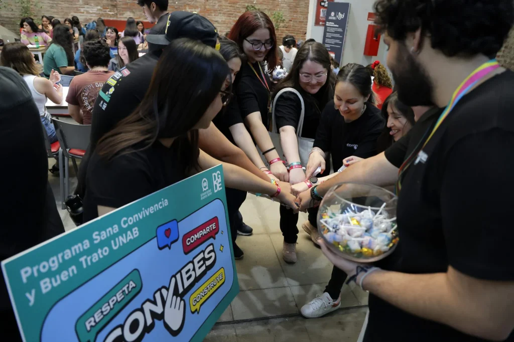 Group of volunteers in black shirts reach across a table to place wristbands on attendees at a community event, with a colorful sign in the foreground.