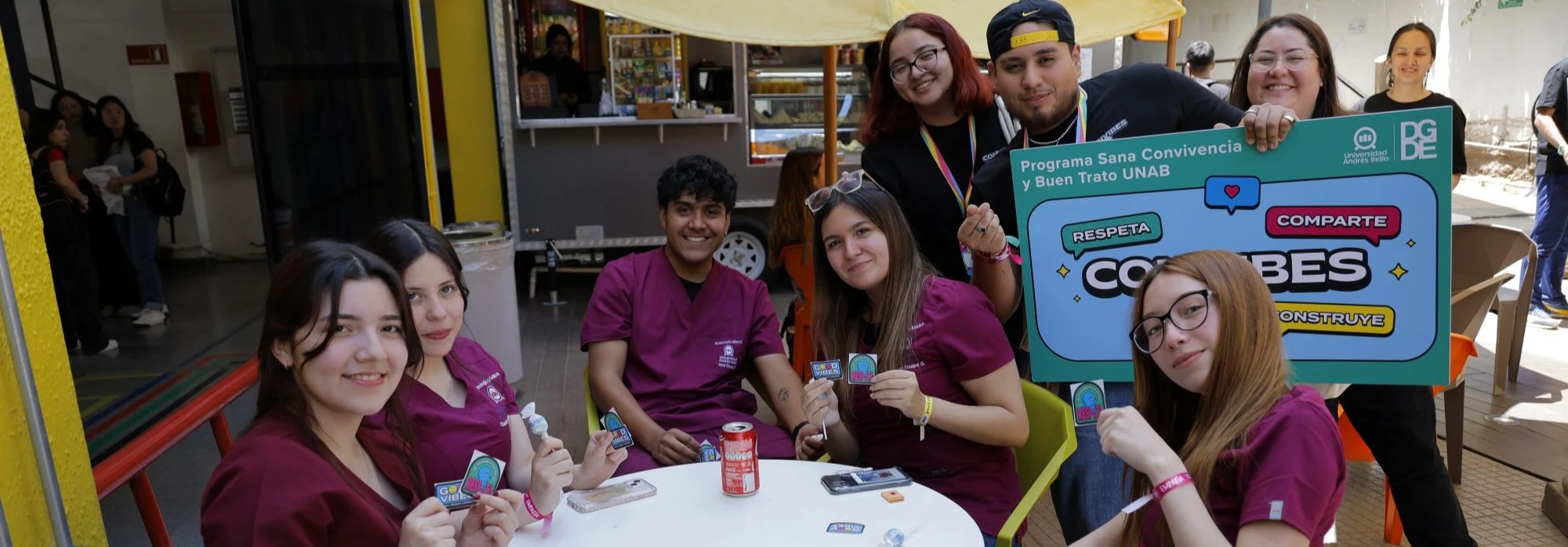 Group of students in maroon scrubs at a booth, smiling and holding badges and lollipops outdoors.
