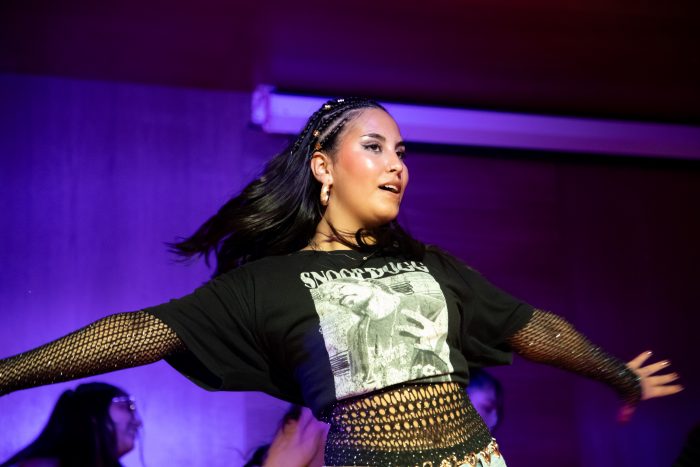 Woman performing on stage with arms outstretched under purple lights, wearing a black graphic crop top and fishnet sleeves.