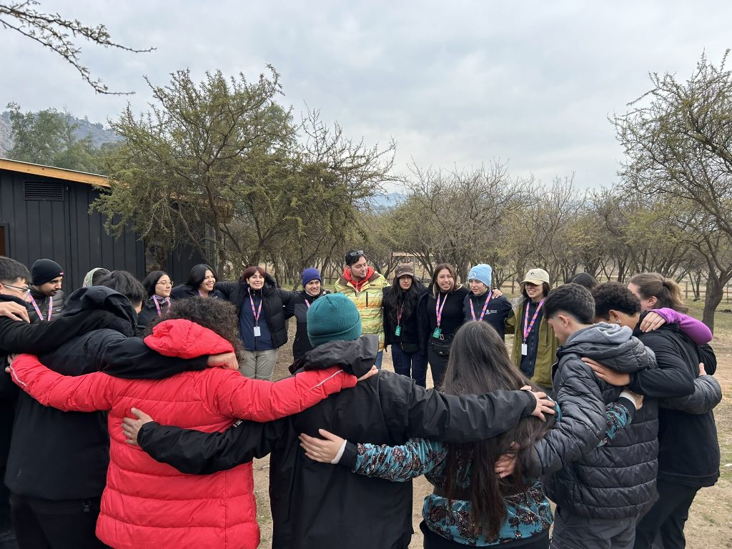 Group of people in winter jackets standing in a circle with arms around each other outdoors on a cloudy day.