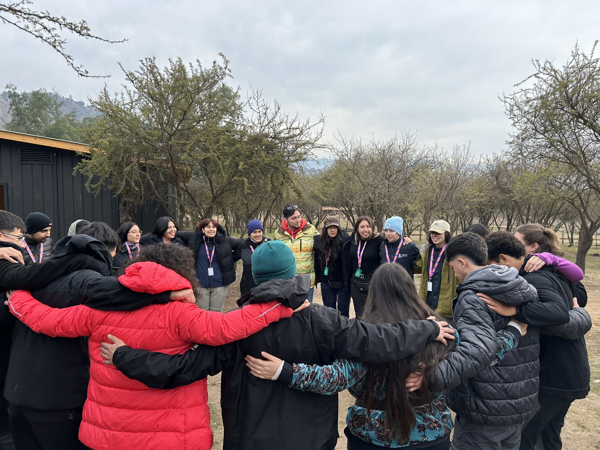 Group of people in winter jackets standing in a circle with arms around each other outdoors on a cloudy day.