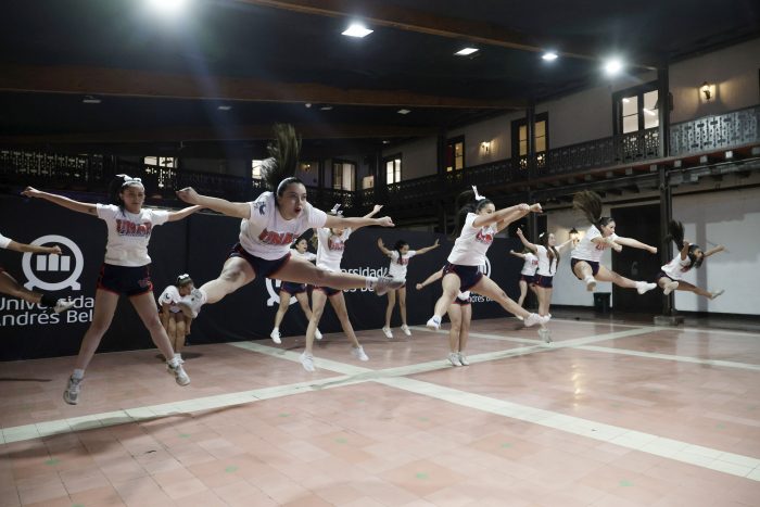 Group of cheerleaders or dancers mid-air jump in a gymnasium during practice, wearing white shirts and shorts on a tiled floor.