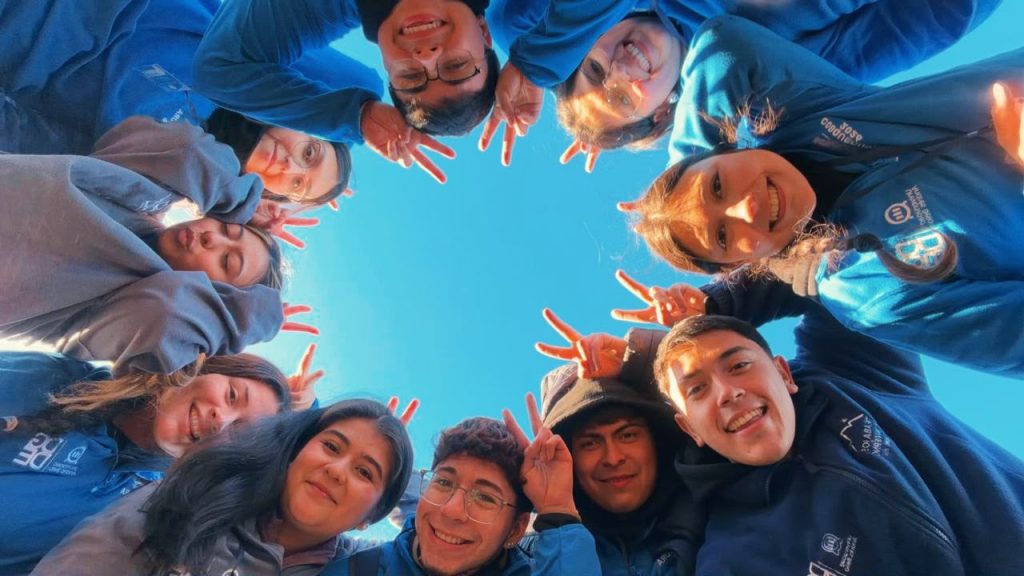 Smiling group of people in blue shirts form a circle, looking down at the camera and making peace signs against a clear blue sky.