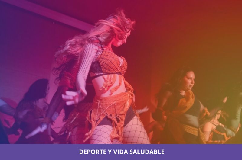 Group of dancers performing on a stage under purple-red lighting, foreground dancer in a mesh top and orange skirt moving energetically.