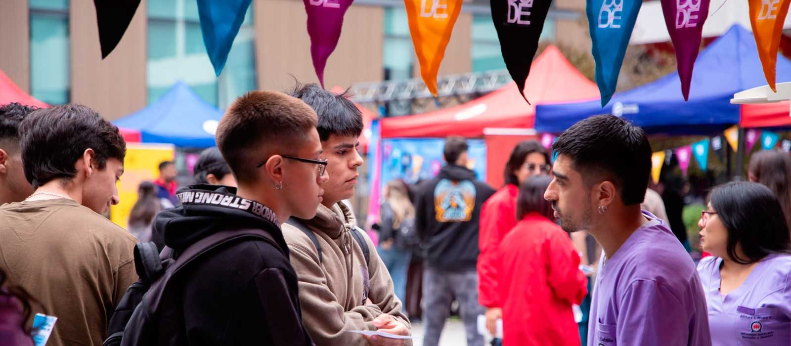 Group of people chatting at an outdoor market or festival with colorful tents and pennant banners above.