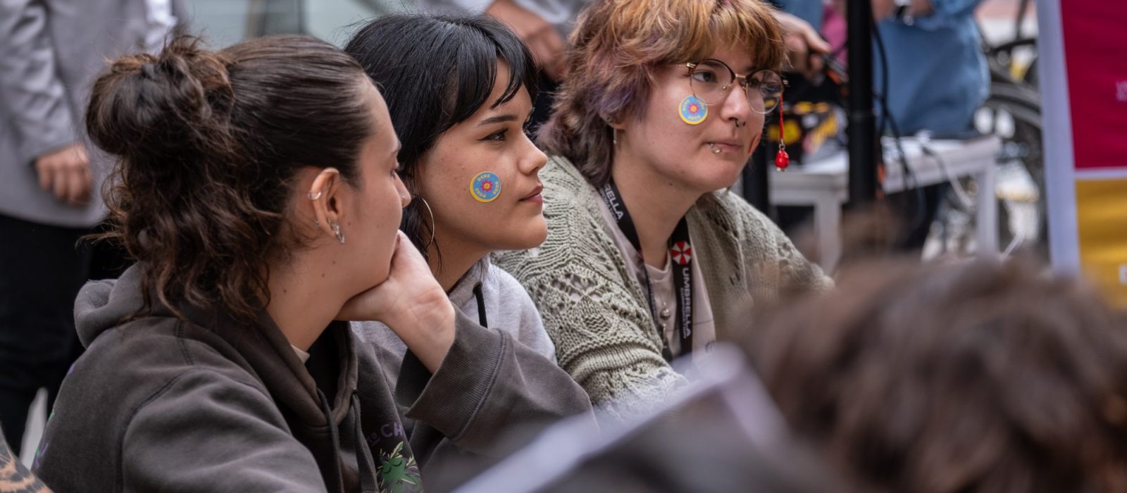 Three young people sit together at an indoor event, two with blue circular stickers on their cheeks and attentive expressions.
