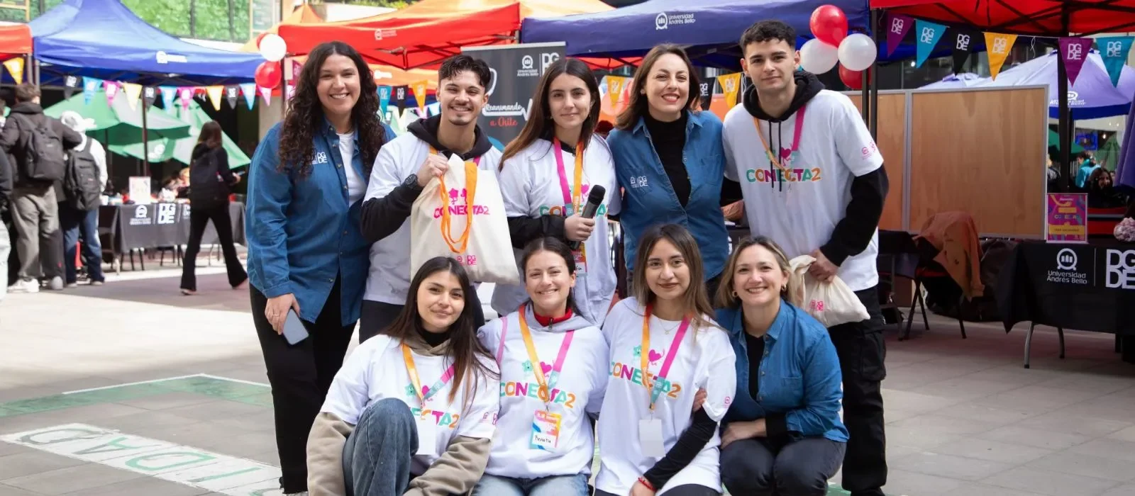 Group of nine volunteers posing and smiling at an outdoor fair with colorful tents, balloons, and banners in the background.