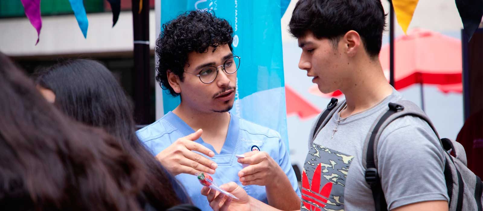 Two young men converse at an outdoor event; one in light blue scrubs with glasses explains a small item to the other with a gray camo shirt and backpack.