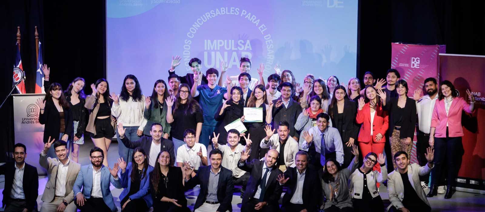Group of diverse students posing on a stage, waving to the camera at a ceremony or presentation event.